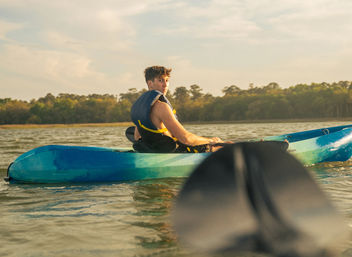 Solo kayaker in a blue kayak wearing a life jacket paddling on a calm lake at golden hour, paddle blade blurred in the foreground and tree-lined shore in the background