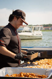 Smiling chef in black uniform and cap grilling shrimp and seasoned chicken on a flat-top at a waterfront marina, with yachts and shoreline buildings in the background