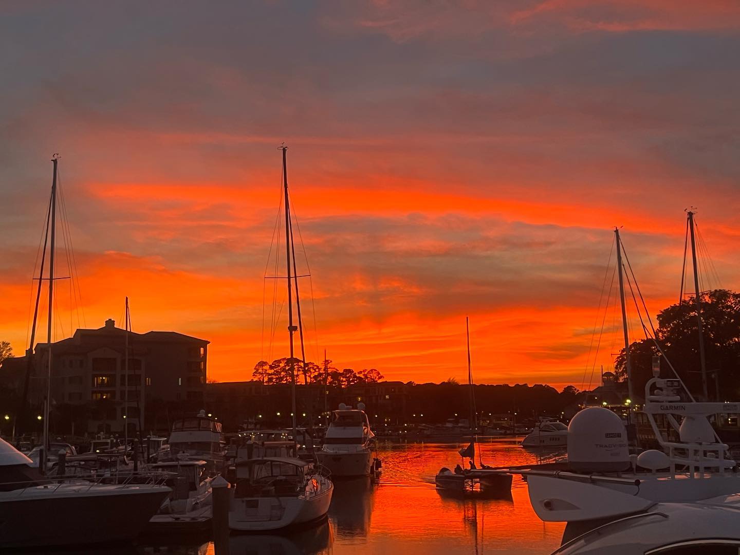Fiery orange-red sunset over a coastal marina with silhouetted sailboat masts and yachts reflecting on calm water, a small dinghy gliding between docks.