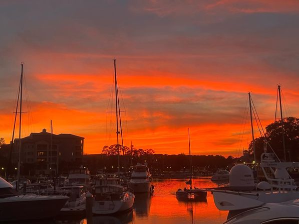 Fiery orange-red sunset over a coastal marina with silhouetted sailboat masts and yachts reflecting on calm water, a small dinghy gliding between docks.