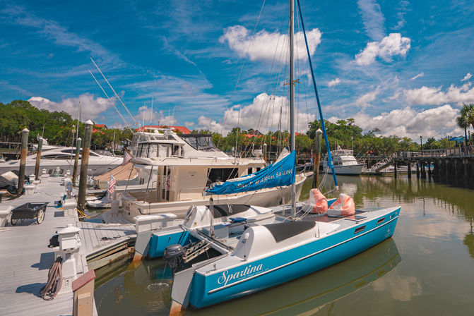 Blue sailboat moored alongside yachts at a sunny coastal marina with wooden docks, palm trees, and puffy clouds reflected in calm water