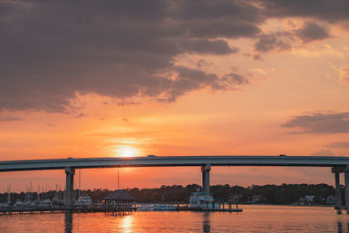 Sunset behind an elevated coastal bridge over a marina, sailboats and docks reflecting warm orange light on calm water.