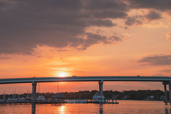 Sunset behind an elevated coastal bridge over a marina, sailboats and docks reflecting warm orange light on calm water.
