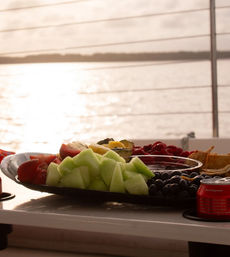 Fresh fruit platter of honeydew, strawberries, grapes and berries on a boat deck table, backlit by a golden sunset over calm water — perfect sunset cruise snack.
