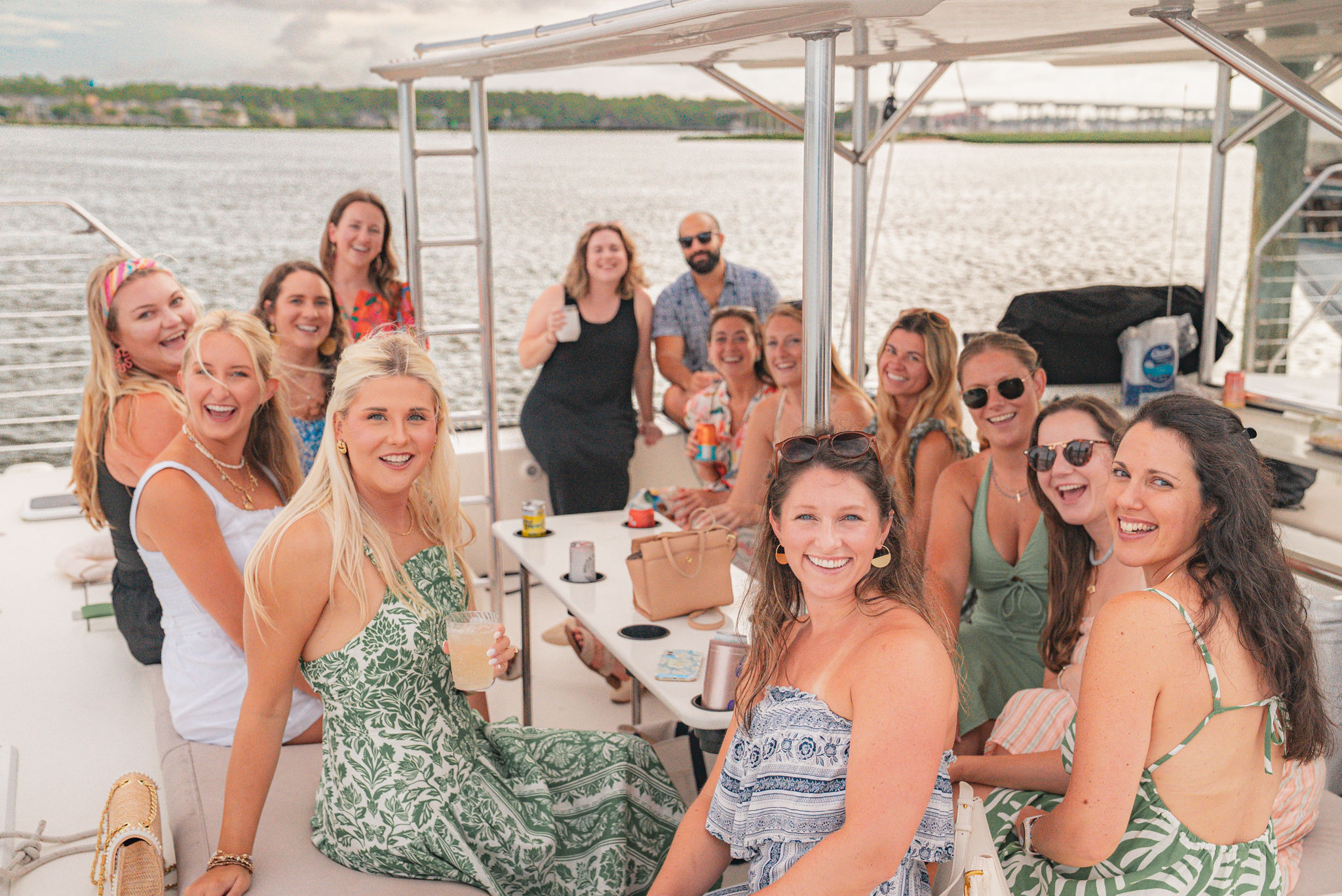 Smiling group of friends on a sunny boat outing in a coastal harbor, gathered under a canopy around a table with cocktails and snacks and docks in the background.