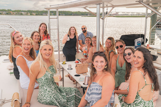 Smiling group of friends on a sunny boat outing in a coastal harbor, gathered under a canopy around a table with cocktails and snacks and docks in the background.