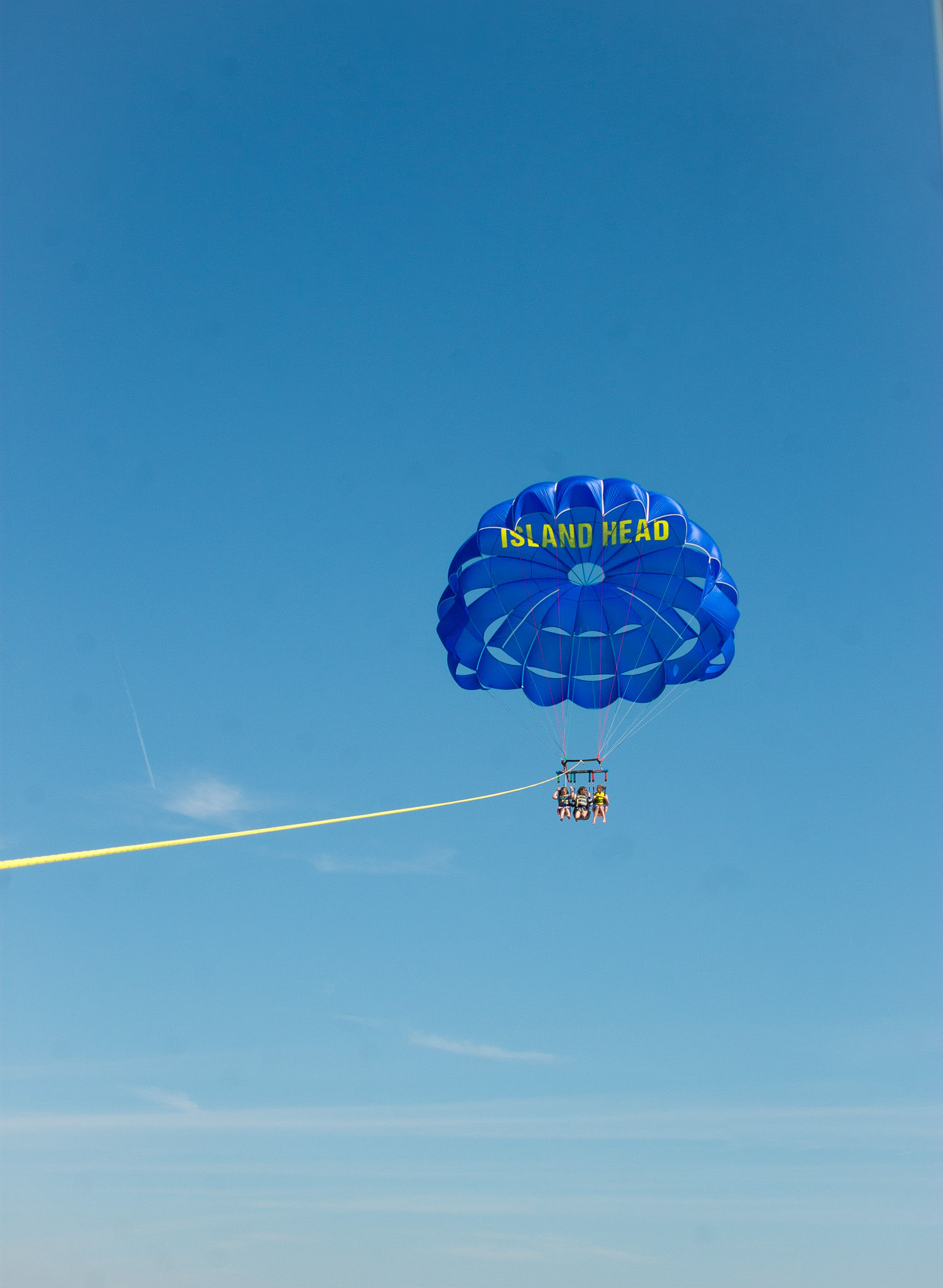 Three people parasailing under a vivid blue parachute over the ocean, tethered by a long yellow tow rope against a clear sunny sky.