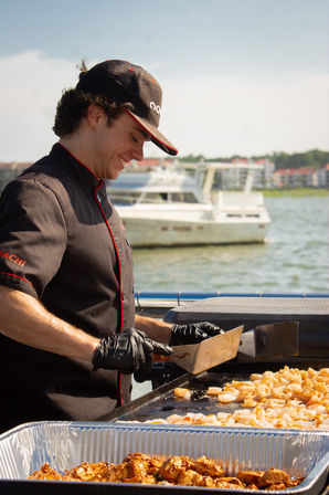 Smiling chef in black uniform and cap grilling shrimp on a flat-top at a waterfront marina with a yacht in the background and an aluminum tray of seasoned grilled bites.