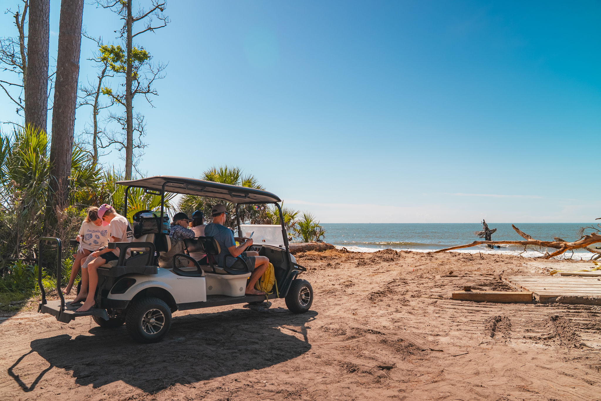 Open-air golf cart with passengers parked on a sandy coastal beach beside palm trees and driftwood, sunny blue sky and calm ocean waves in the background.