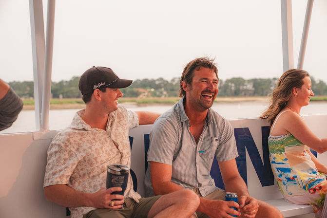 Three friends laughing on a summer boat ride near a tree-lined coastal shoreline at golden hour, sitting on a bench and holding drinks.