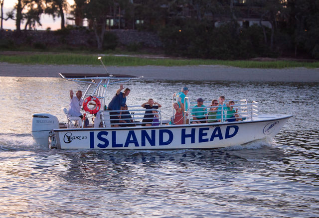 White passenger tour boat with large blue lettering cruising a calm coastal inlet at dusk, cheerful people waving from the deck, lifebuoy on the rail and rippling water reflections