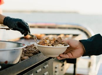 Hand-held rice bowl topped with grilled beef and sesame seeds being served with tongs from a gloved cook at an outdoor waterfront grill, street-food style.