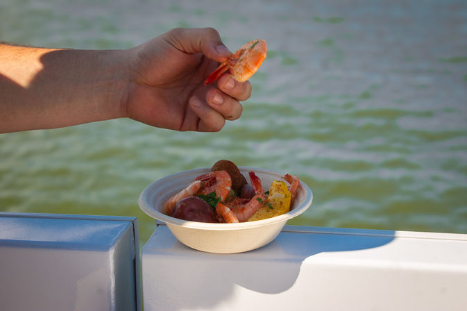 Hand holding a seasoned shrimp above a bowl of seafood boil (shrimp, corn, potatoes) resting on a boat railing with green water in the background