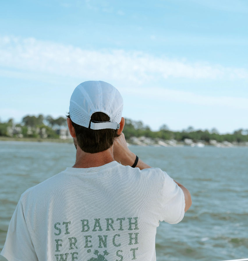 Rear view of a person in a white cap and 'St Barth' t-shirt on a boat, gazing across a calm bay toward a tree-lined shoreline and bright blue sky.
