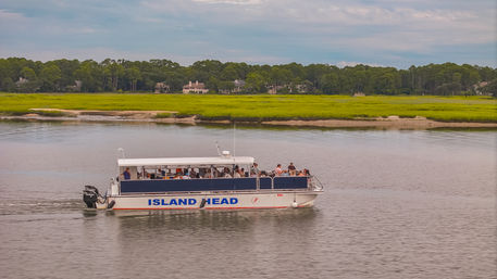 Passenger tour boat cruising on a calm coastal river past bright green salt marsh and tree-lined waterfront homes under a cloudy sky.