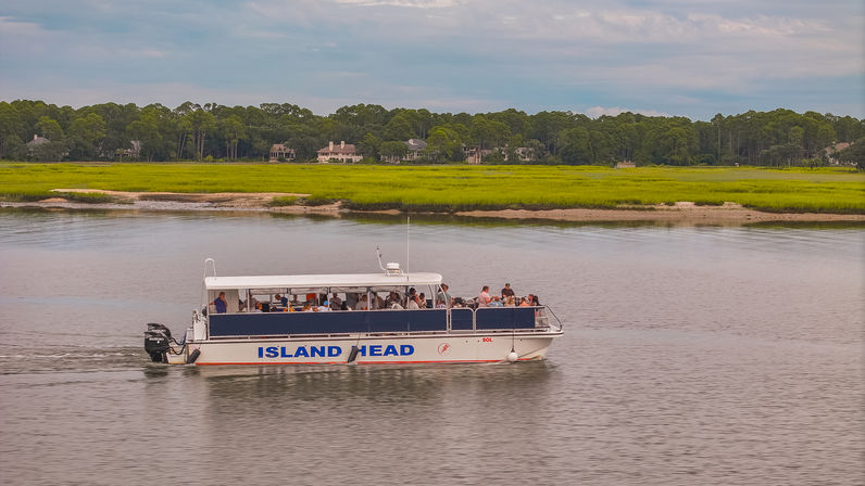Passenger tour boat cruising on a calm coastal river past bright green salt marsh and tree-lined waterfront homes under a cloudy sky.