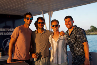 Four people smiling on a boat during golden-hour sunset, wearing sunglasses and casual summer shirts while relaxing on a sunset cruise with water and a tree-lined shoreline in the background.