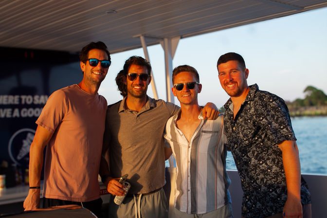 Four people smiling on a boat during golden-hour sunset, wearing sunglasses and casual summer shirts while relaxing on a sunset cruise with water and a tree-lined shoreline in the background.