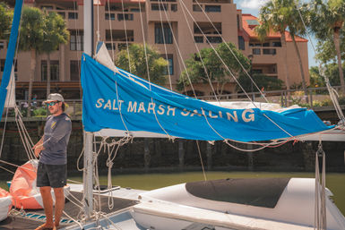 Sunny coastal marina scene with a person on a sailboat, bright blue sail cover with white lettering, rigging and inflatable dinghy, palm trees and waterfront buildings in the background.