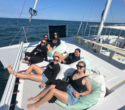 Group of friends lounging on a catamaran trampoline and mint cushions, wearing sunglasses and summer clothes, enjoying a sunny sailing day on the calm blue ocean.