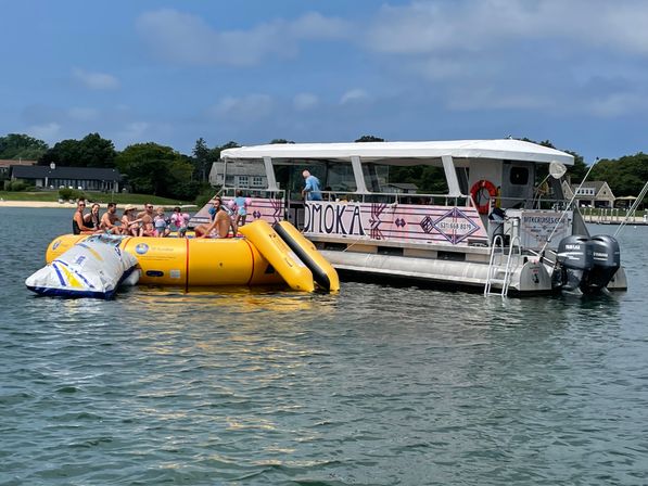 Sunny pontoon party in a coastal bay with people lounging on a boat and a bright yellow inflatable water trampoline/slide tethered beside it, shoreline homes in the background.