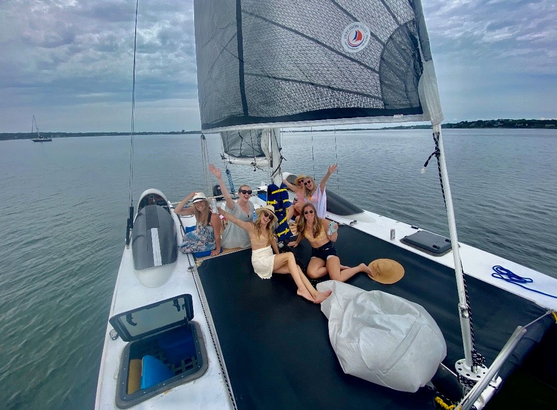 Group of friends in summer outfits and sun hats relaxing and waving on the trampoline deck of a catamaran sailboat in a calm coastal bay under a cloudy sky