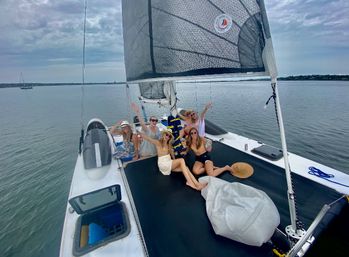 Group of friends in summer outfits and sun hats relaxing and waving on the trampoline deck of a catamaran sailboat in a calm coastal bay under a cloudy sky