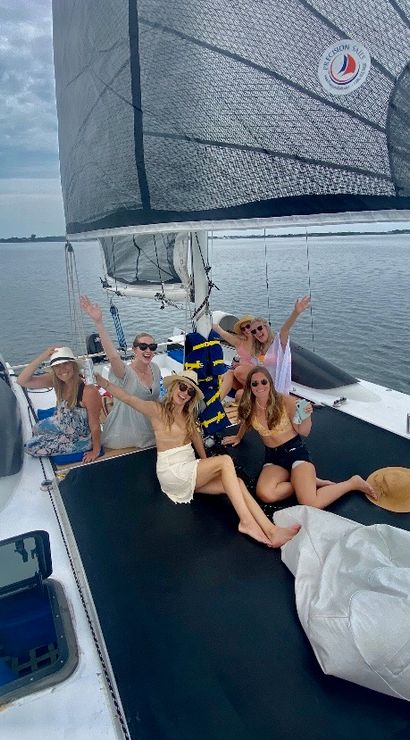Group of friends in summer outfits and sun hats relaxing and waving on the trampoline deck of a catamaran sailboat in a calm coastal bay under a cloudy sky