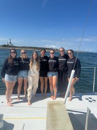 Seven friends in matching 'BRIDESMAID' and 'MAID OF HONOR' shirts smiling barefoot on a sailboat deck with a lighthouse and rocky shoreline under a bright blue sky — bachelorette boat outing on coastal waters.