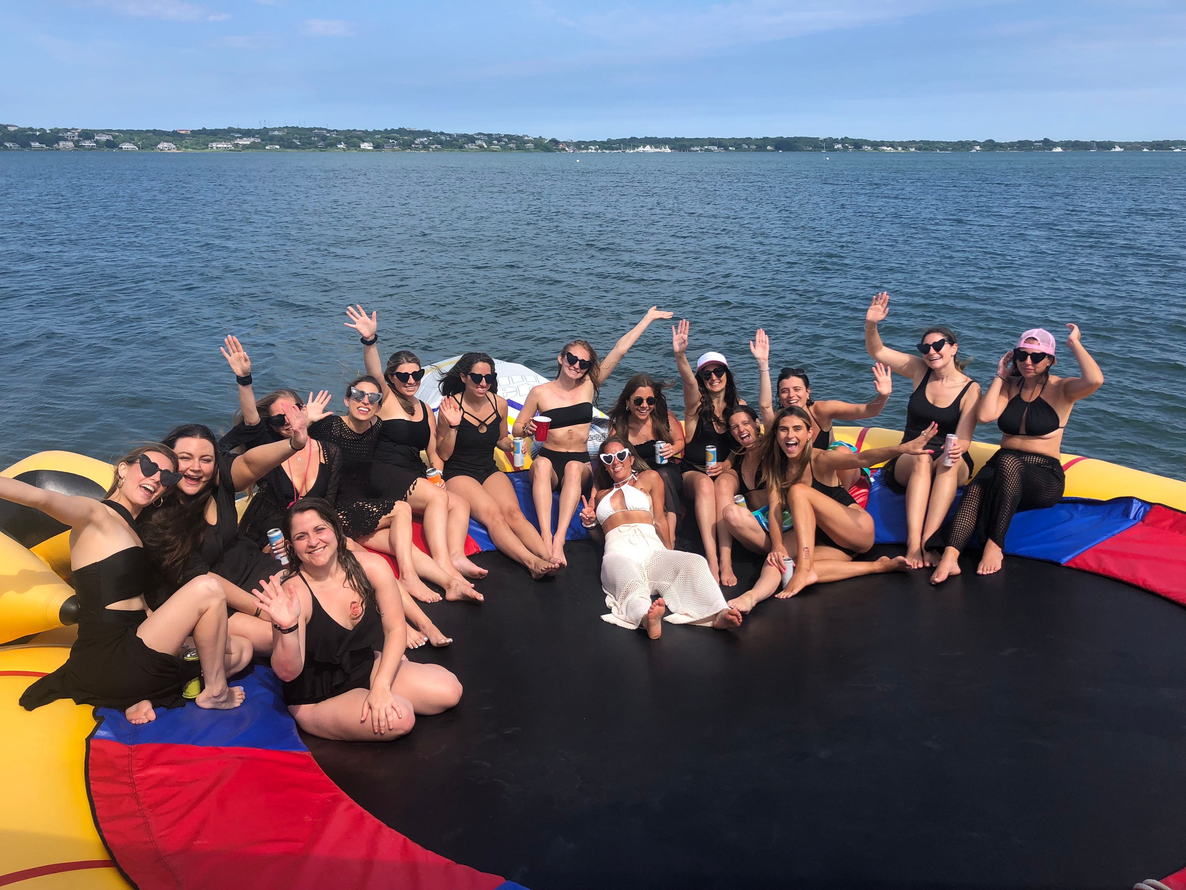 Group of friends in swimsuits smiling and waving on a colorful inflatable water trampoline anchored on a sunny lake with shoreline and blue sky in the background