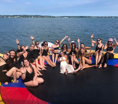 Group of friends in swimsuits smiling and waving on a colorful inflatable water trampoline anchored on a sunny lake with shoreline and blue sky in the background
