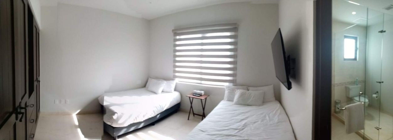 Bright minimalist guest bedroom with two twin beds and white linens, striped roller blinds, wall-mounted TV and glass-enclosed ensuite bathroom.
