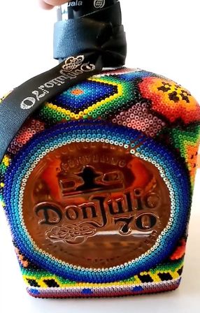 Amber tequila bottle with embossed medallion, covered in vibrant Huichol-style beaded geometric patterns and a black ribbon around the neck