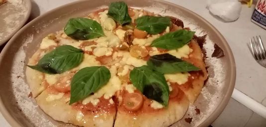 Fresh margherita pizza cut into slices with gooey mozzarella, cherry tomato slices and glossy basil leaves on a round ceramic plate at a casual dining table.