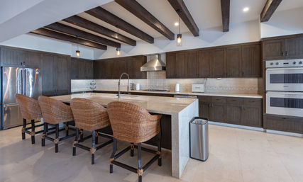 Open-concept luxury kitchen with a large stone island, four woven rattan bar stools, dark wood cabinets, exposed wood ceiling beams, pendant lights, stainless steel refrigerator and double wall ovens with tiled backsplash