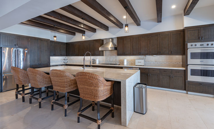 Open-concept luxury kitchen with a large stone island, four woven rattan bar stools, dark wood cabinets, exposed wood ceiling beams, pendant lights, stainless steel refrigerator and double wall ovens with tiled backsplash