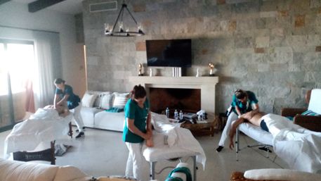 Three massage therapists in teal uniforms working on clients on portable massage tables in a bright, living-room-style spa with a stone accent wall, fireplace, wall-mounted TV and large window