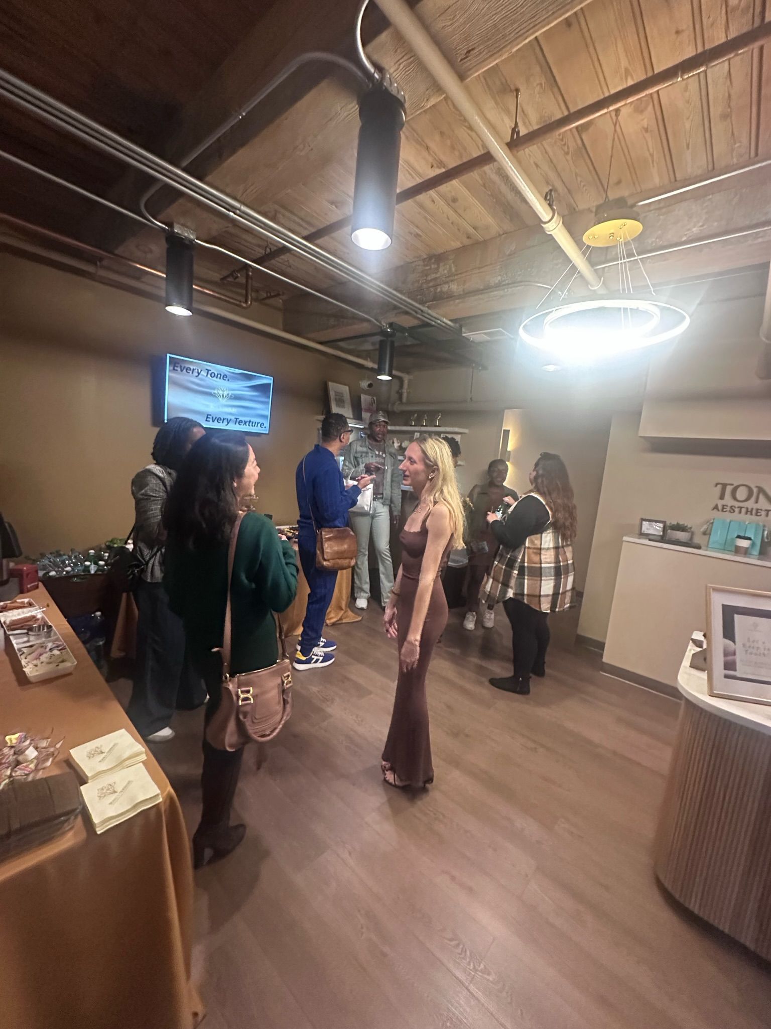 Guests mingling at a casual networking reception in a loft-style interior with exposed wooden ceiling beams, circular pendant light, wood floors, snack table and reception desk.