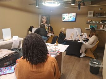 Group of people in a cozy indoor studio doing a casual art workshop with easels, magazines and craft supplies on tables under pendant lights and a wall-mounted TV.