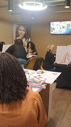 Group of people in a cozy indoor studio doing a casual art workshop with easels, magazines and craft supplies on tables under pendant lights and a wall-mounted TV.