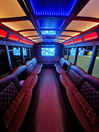 Luxury party bus interior with red and blue LED ceiling lights, quilted leather bench seating along both sides, hardwood aisle, and multiple flat-screen TVs.