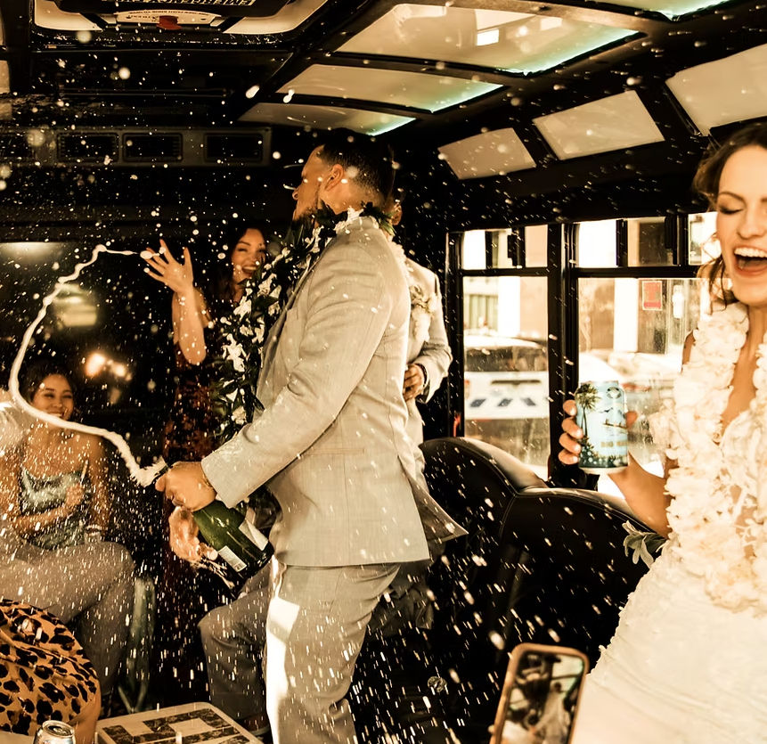 Wedding party celebrating inside a city party bus, champagne spraying as a man in a suit pops the bottle and guests, including a laughing bride in white and friends with leis, cheer amid bubbly foam.