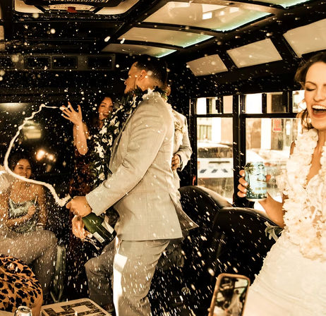 Wedding party celebrating inside a city party bus, champagne spraying as a man in a suit pops the bottle and guests, including a laughing bride in white and friends with leis, cheer amid bubbly foam.