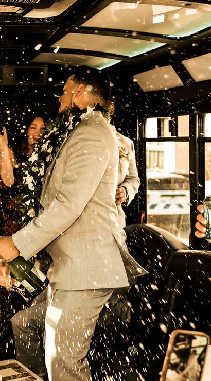 Wedding party celebrating inside a city party bus, champagne spraying as a man in a suit pops the bottle and guests, including a laughing bride in white and friends with leis, cheer amid bubbly foam.