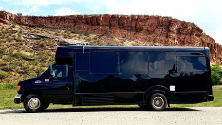 Sleek black shuttle bus parked by red rock cliffs and grassy canyon under a bright blue sky