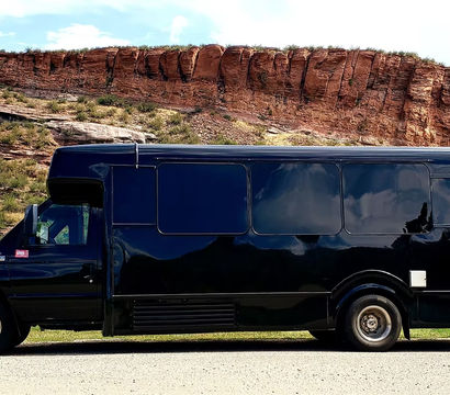 Sleek black shuttle bus parked by red rock cliffs and grassy canyon under a bright blue sky