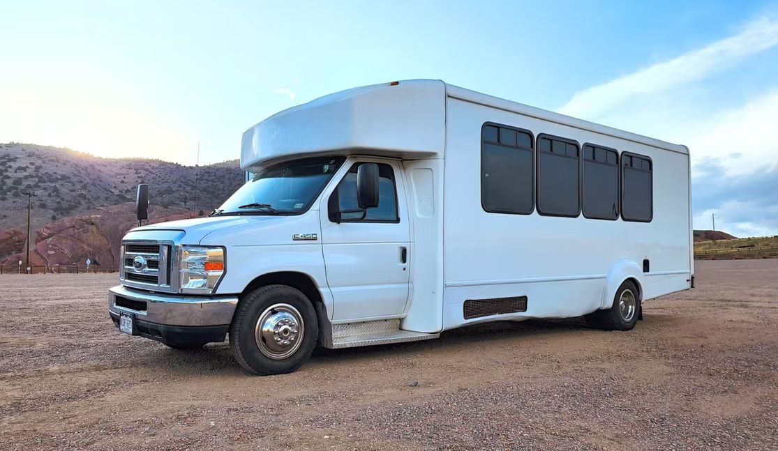White shuttle bus (Ford E-450 chassis) parked on a gravel lot with red rock hills and a pastel sunset sky — side view showing tinted passenger windows.