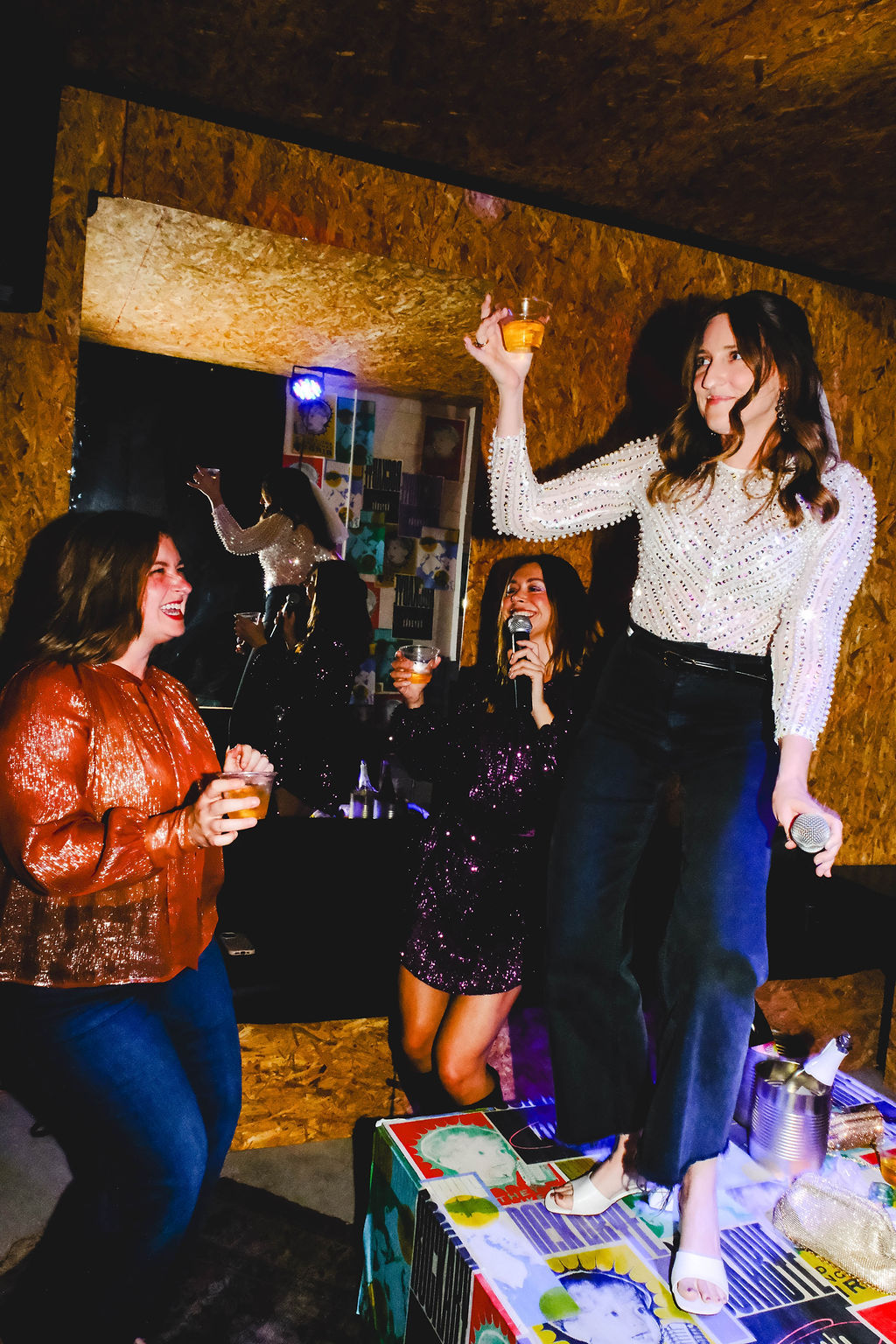 Women at an indoor karaoke night, one standing on a table with a microphone and drink while friends cheer with cocktails.