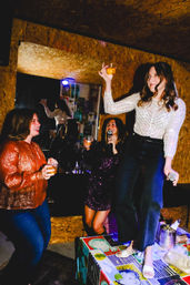 Women at an indoor karaoke night, one standing on a table with a microphone and drink while friends cheer with cocktails.
