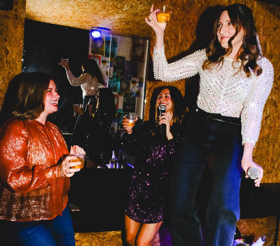 Women at an indoor karaoke night, one standing on a table with a microphone and drink while friends cheer with cocktails.